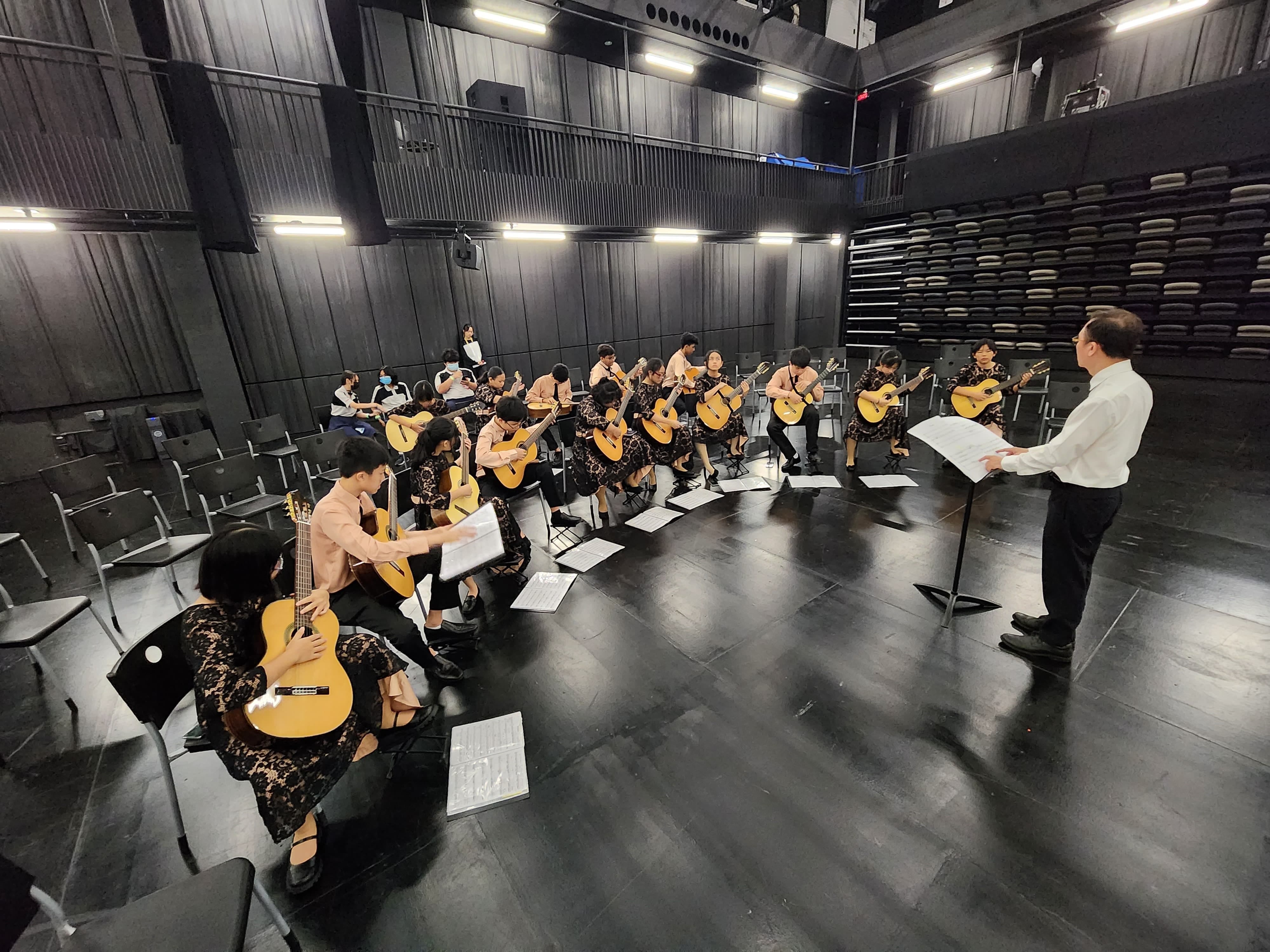A conductor leads a large group of classical guitar players rehearsing on a black stage in an empty theater.